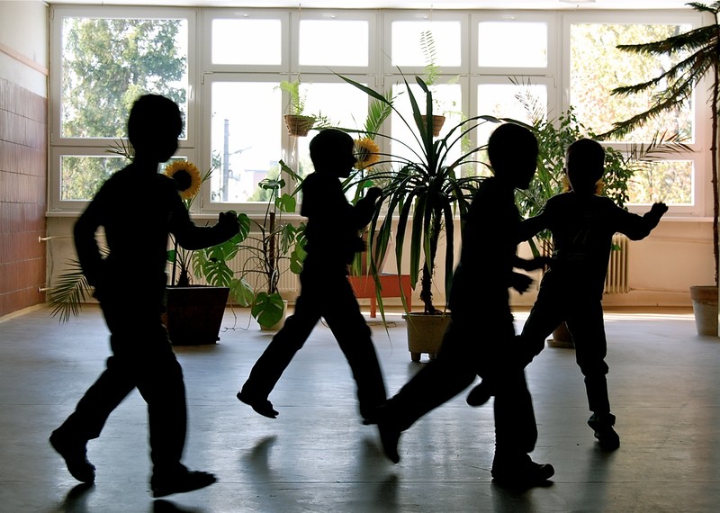 Silhouettes of children playing in a large school room with plants in the background.