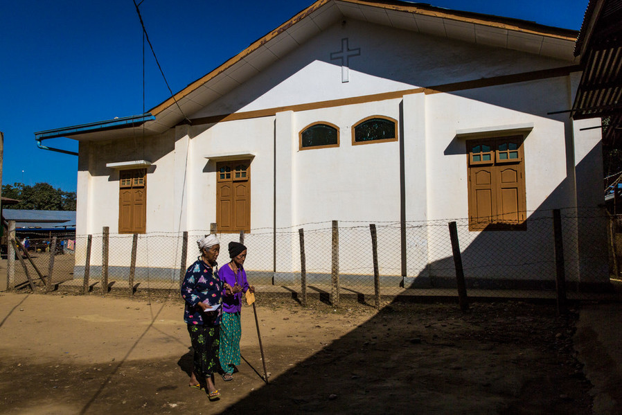 two older women walk past a white building on their way to see a health worker at an IDP camp. One of the women is walking with a walking stick. 