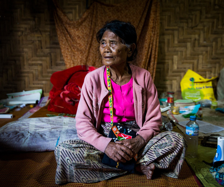 a portrait of an older woman sits cross-legged in her shelter in Kachin State Myanmar. She is wearing a bright pink shirt and cardigan.