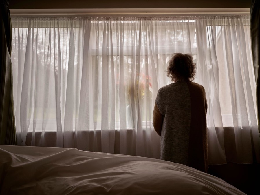 An older woman looks out of a window with curtains while self isolating during the Covid-19 pandemic.