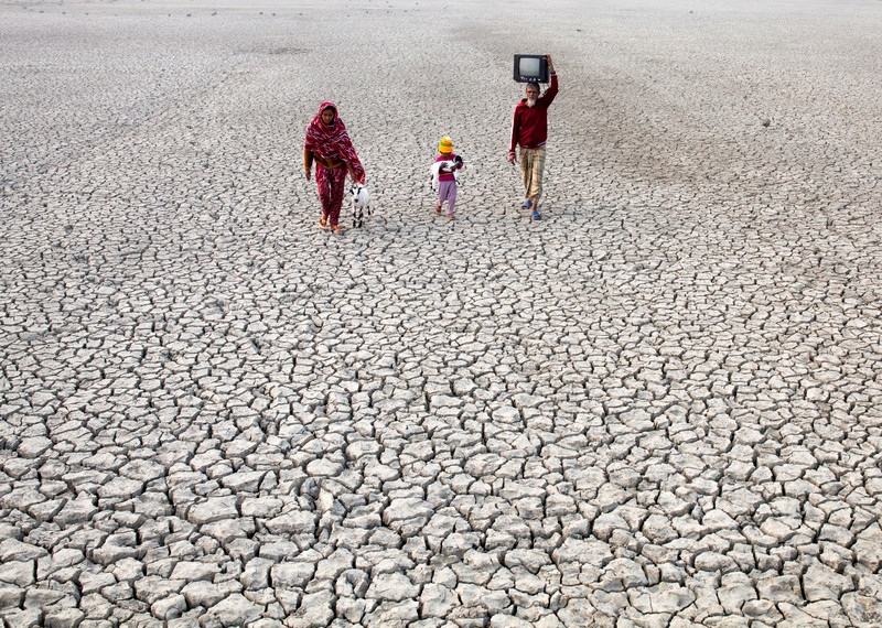 A family walks across a dried river bed. The ground is made up of deeply cracked, white soil that continues past the horizon. The family includes a woman wearing pink fabric that drapes over her head, a young child carrying a small white goat and a man wearing a red jumper and carrying a television set. 