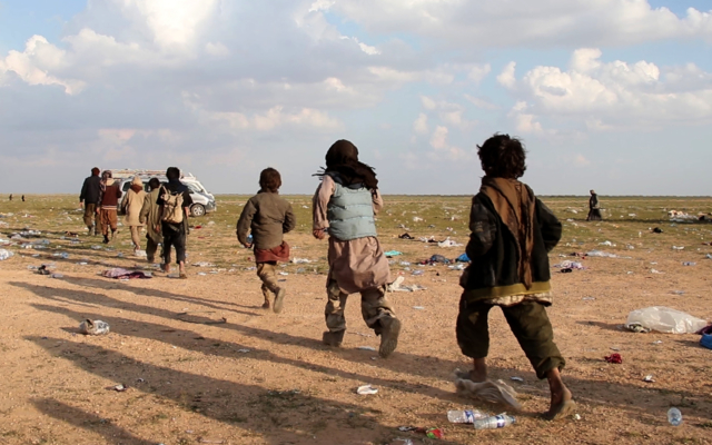 a group of boys walk in a row towards a vehicle. The ground is covered in litter and bottles. 