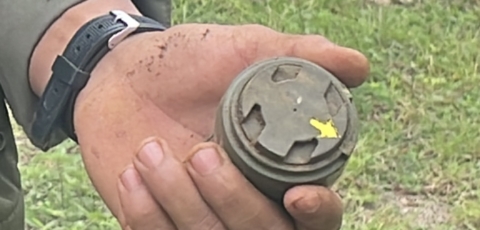 a close-up show of a hand holding an M-14 antipersonnel landmine. It is small, about the size of the person's palm and dark green. 