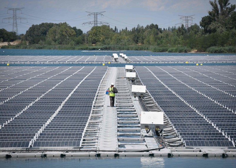 a worker in a white helmet and a high visibility jacket walks amid rows of solar panels. 
