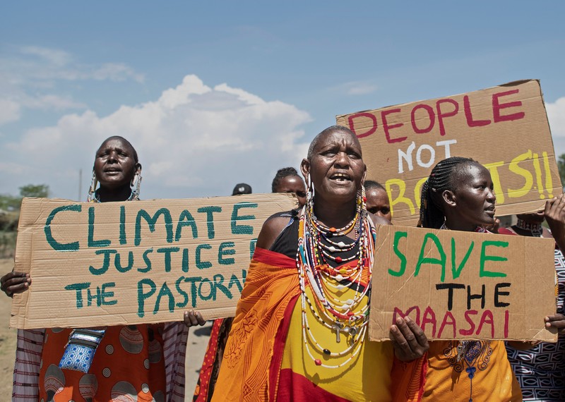 a group of women in traditional Masai dress hold up signs that read "Save the Masai" and "People not profits"