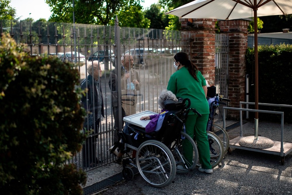 A woman in a wheel chair and a care worker stand at the gate of a care home, talking to people outside. There is a protective barrier on the gate to avoid the spread of the virus.