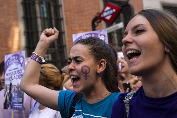 two women are in focus in the image, both look as though they are speaking or chanting. The one on the left has a Venus symbol painted in purpose on her cheek and is holding up a fist. 